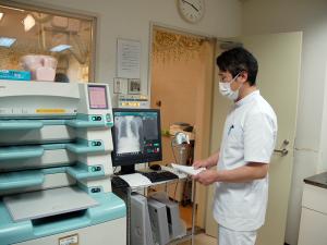 壁に時計が掛けられた部屋で、白衣を着た診療放射線技師の男性がモニターに映し出された画像を見ている写真