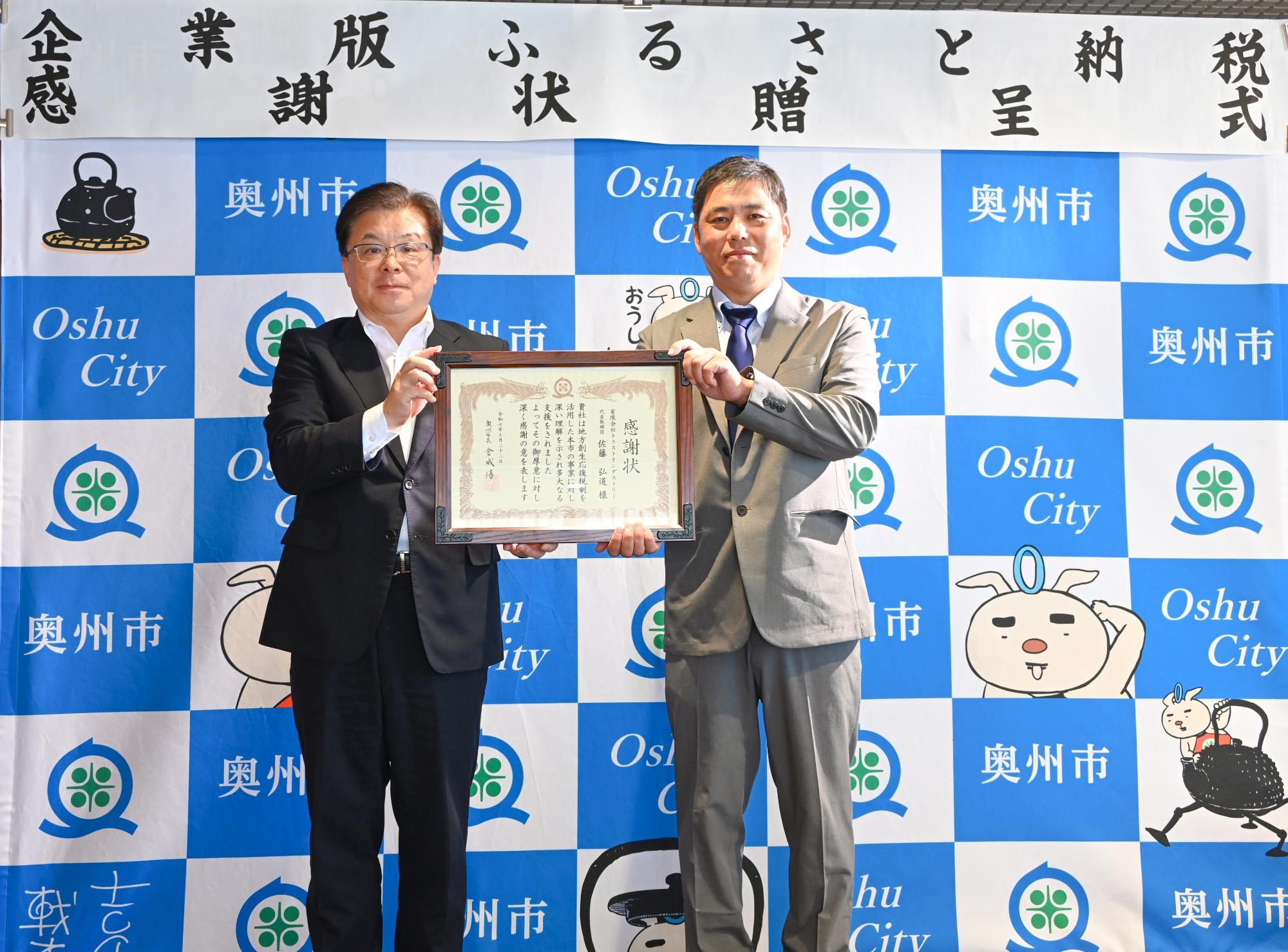 「企業版ふるさと納税感謝状贈呈式」の横断幕が掲げられた奥州市のバックボードの前で2人で感謝状を持って立つ倉成市長と佐藤代表取締役の写真