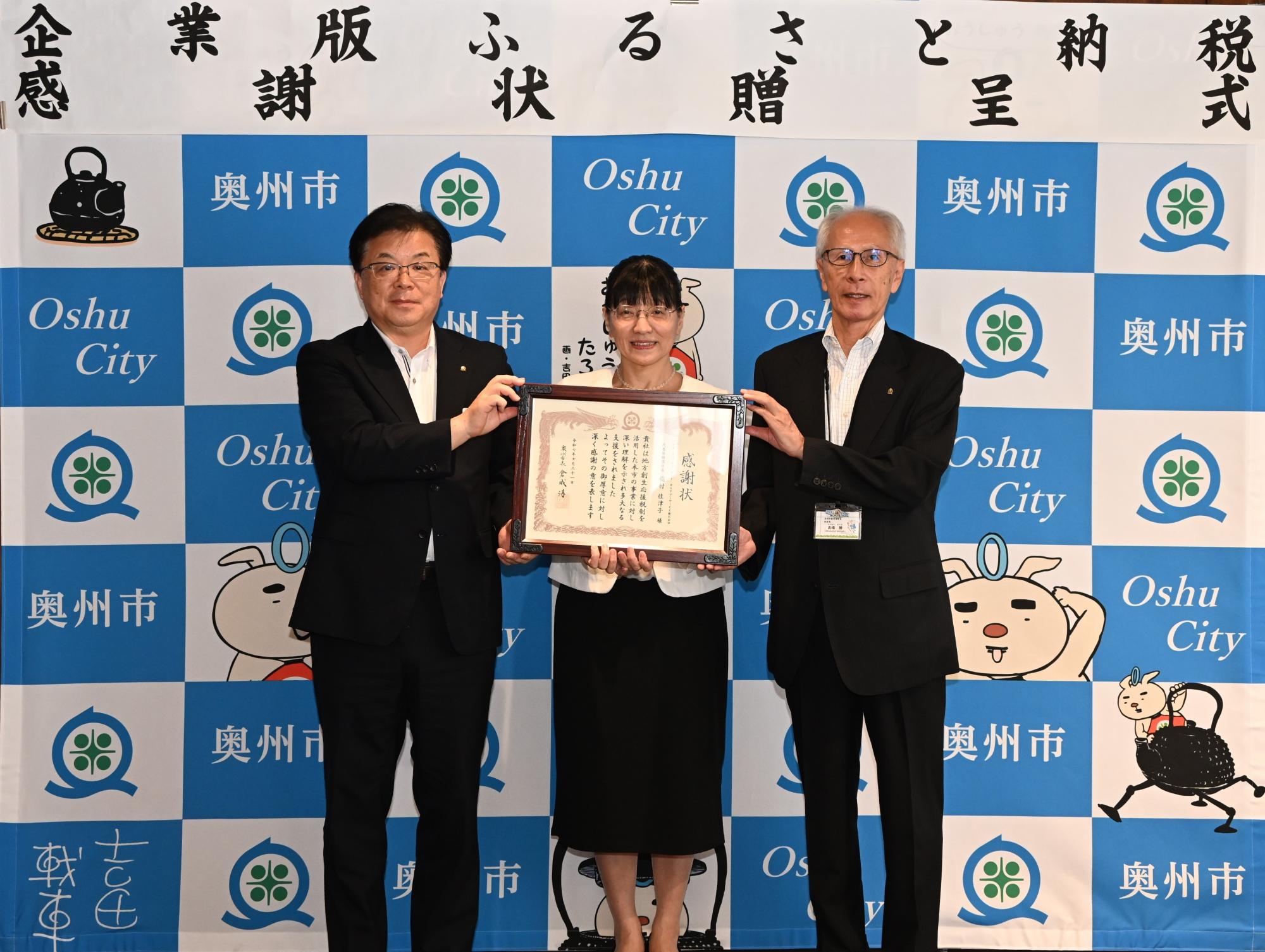 「企業版ふるさと納税感謝状贈呈式」の横断幕が掲げられた奥州市のバックボードの前で3人で感謝状を持って立つ倉成市長、稲村代表取締役社長、高橋教育長の写真