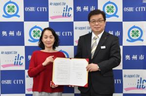 赤色の服を着た女性とスーツ姿の男性が協定書を一緒に持って笑顔で写っている写真