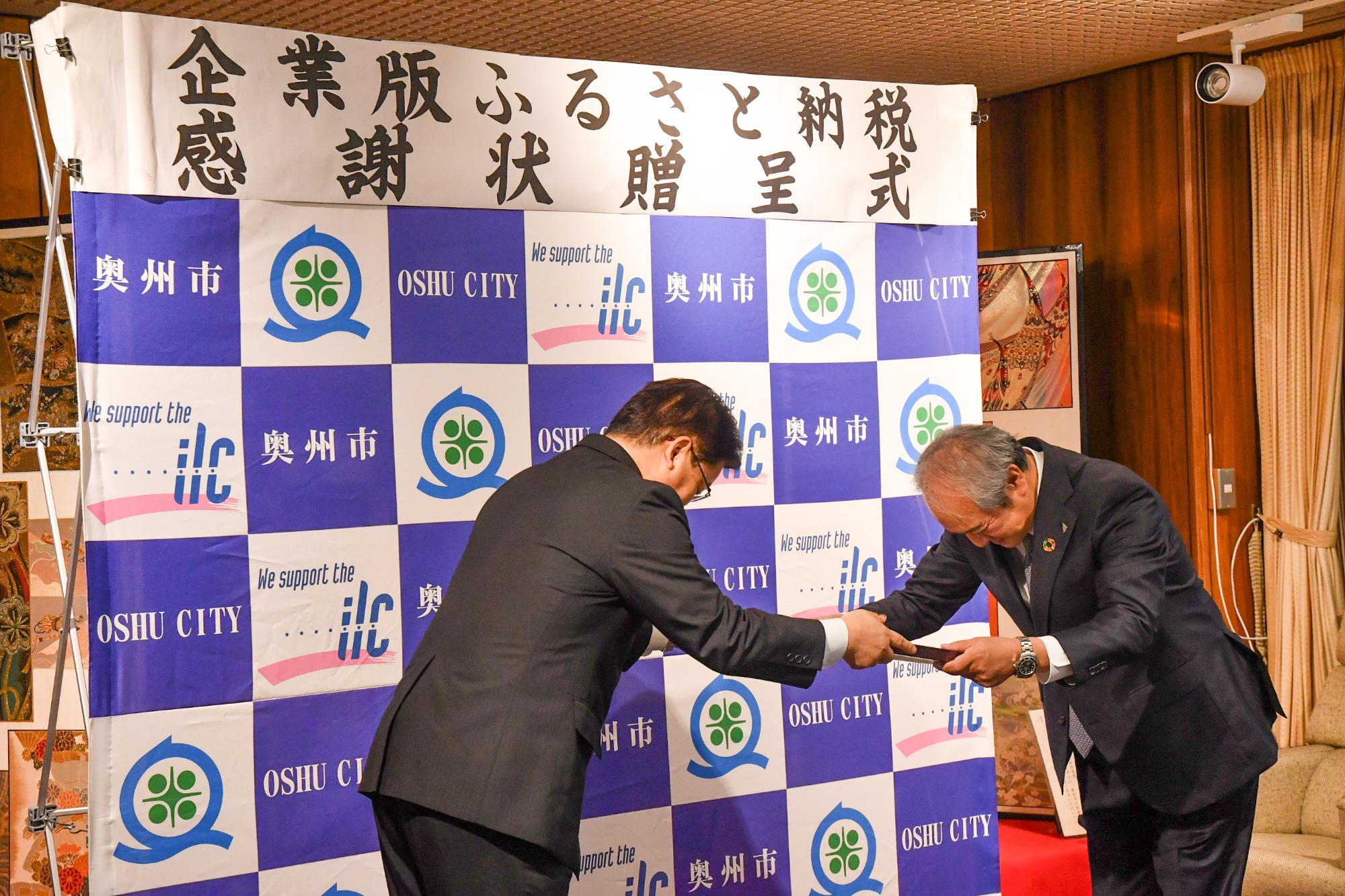 「企業版ふるさと納税感謝状贈呈式」の横断幕が掲げられた奥州市のバックボードの前で中川専務取締役に感謝状を手渡す倉成市長の写真