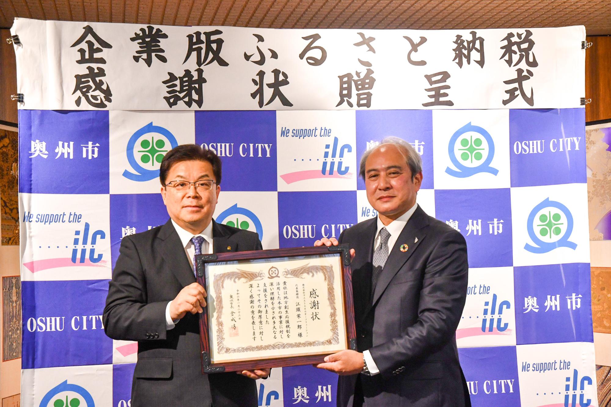「企業版ふるさと納税感謝状贈呈式」の横断幕が掲げられた奥州市のバックボードの前で2人で感謝状を持って立つ倉成市長と中川専務取締役の写真