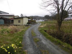 砂利道の左側に民家が建ち、右側に畑が広がっている奥に向かってカーブしている細い道の写真