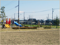 更地の向こう側に青や黄色の滑り台のある公園や、住宅街が広がっている写真