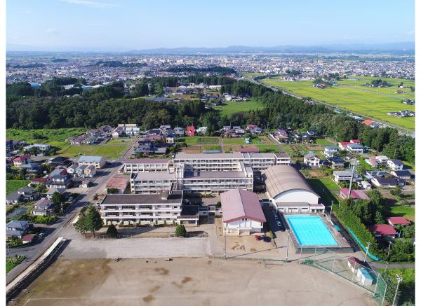 校庭の先に建てられた校舎や体育館、プールなどの建物の全貌を、後ろに広がる街並みと一緒に上空から写した奥州市立水沢南中学校の外観写真