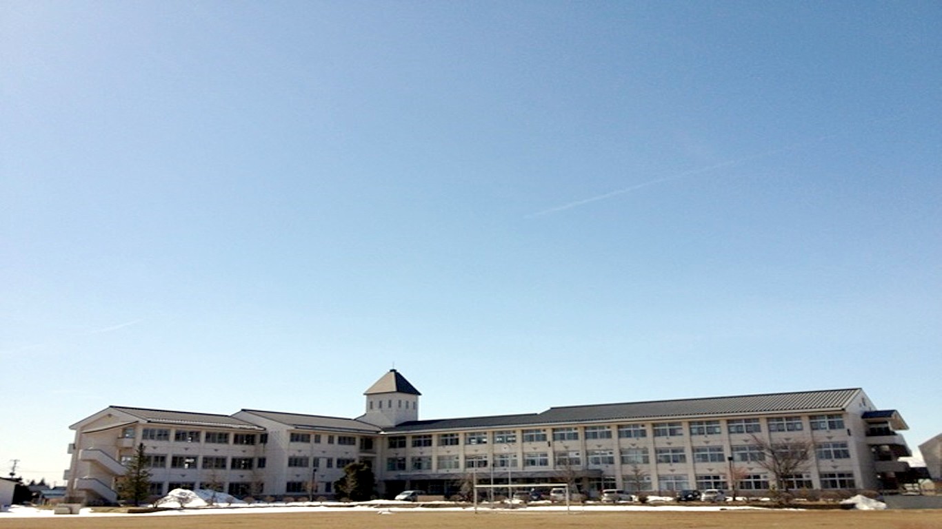 所々に雪が残る校庭の先に3階建てでL字型になっている校舎が建っており、校舎の中心部分には方形屋根のついた小さな建物が突き出している奥州市立江刺第一中学校の外観写真