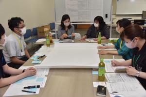 テーブルの上に広げた白紙の模造紙と手元の資料を見ながら話し合いをしているグループの写真