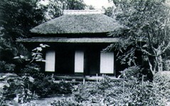 木々に囲まれた中に萱葺き屋根の建物が建っている白黒写真