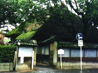 苔のはえた大きな三角の茅葺屋根の建物が瓦のついた塀や生垣に囲まれており、門の前には白い木の立て看板がある咸宜園の写真