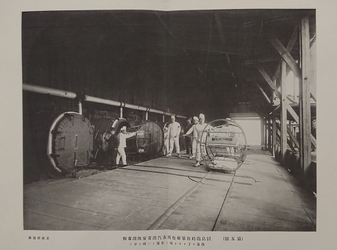 木造の天井の高い工場内で、パイプの通った大きな樽のようなものが左側に並んでおり、白い上下の服を着た人々が作業をしている様子の写真