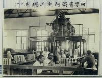 工場内の後方に大きな機械が置かれ、手前の机に座った人々が作業をしている工場内の白黒写真