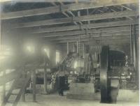 天井に木の柱が横に並んでおり、工場内には大きな車輪のような機械が置かれている工場内の白黒写真