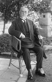 屋外で椅子に座り、帽子を膝に置き、スーツ姿で東京市長時代の後藤新平氏の白黒写真
