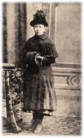 帽子を被り、両端の開いた円筒形の手袋に両手を入れて記念撮影をしている春子夫人の少女時代の白黒写真
