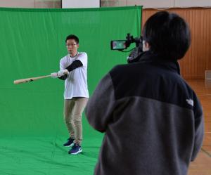 右側の男性が、緑色のスクリーンの上に立っている秦和弘助教がバットをスイングしている様子を撮影している写真