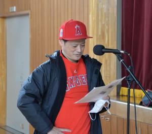 赤色のキャップにシャツを着た本間教授が左手に用紙を持ち、スタンドマイクの前に立って音声収録を行っている様子の写真