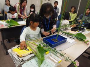 青いトレーの中の剣山に花が挿してあり、指導者の元、女の子が華道を習っている写真