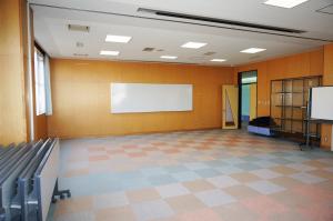 壁にホワイトボードが設置され、部屋の端に畳んだ長机などが置かれた、オレンジとグレーの四角のパターンのカーペット敷きの会議室の写真