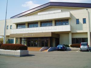 白い外壁で入り口にあがるオレンジ色の階段とスロープがあり、駐車場に2台の車が停まっている丹沢総合体育館の外観写真
