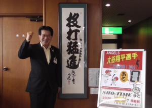 開幕カウントダウンのパネルと「投打猛進」と書かれた掛け軸の横でガッツポーズ をしている小沢昌記奥州市長の写真