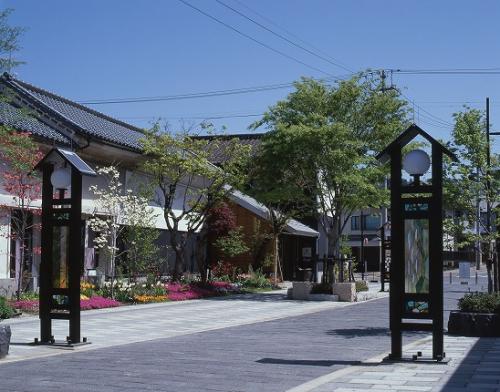 蔵風・和風の建物が立ち並び、建物の横にはピンク・白・黄色の花々や木々が植えられており、通路の両端にステンドグラスの装飾が施された街灯が立っている蔵まちの写真