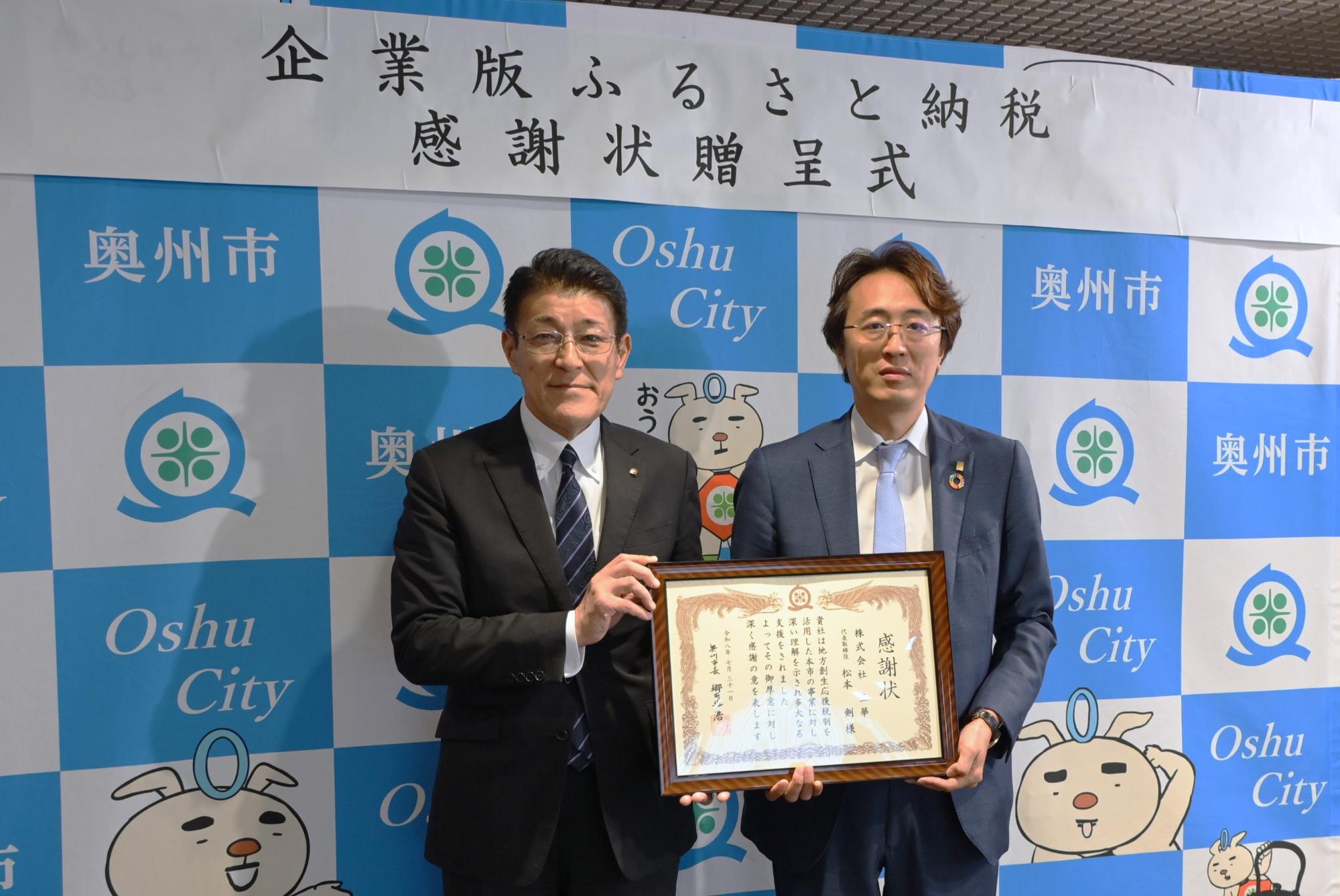 「企業版ふるさと納税感謝状贈呈式」の横断幕が掲げられた奥州市のバックボードの前で松本代表取締役と郷右近市長が並んで感謝状を手に持つ写真