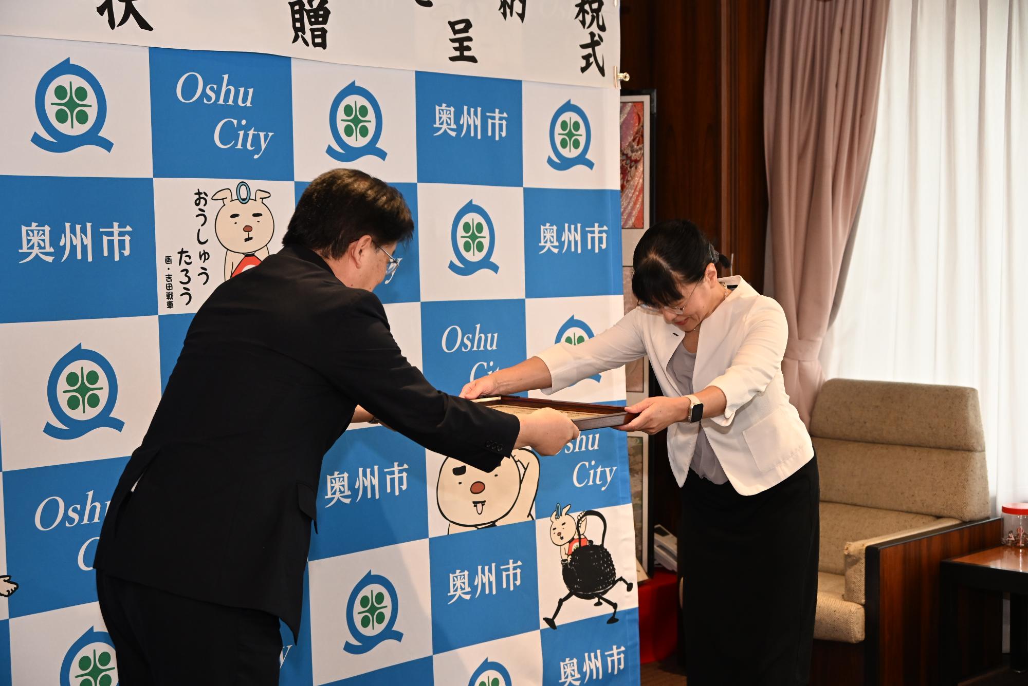 「企業版ふるさと納税感謝状贈呈式」の横断幕が掲げられた奥州市のバックボードの前で稲村代表取締役社長に感謝状を手渡す倉成市長の写真
