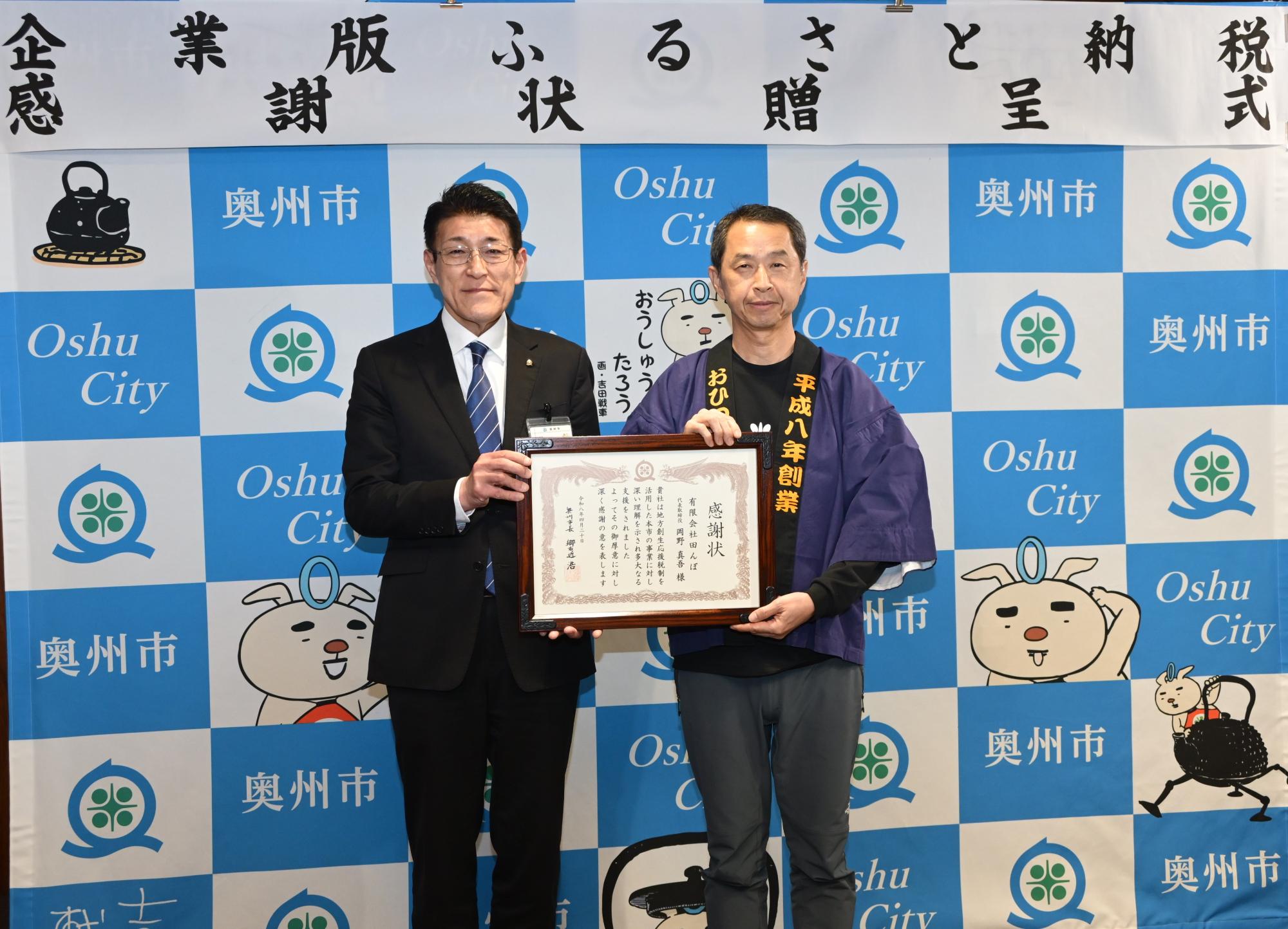 「企業版ふるさと納税感謝状贈呈式」の横断幕が掲げられた奥州市のバックボードの前で岡野代表取締役に感謝状を手渡す郷右近市長の写真