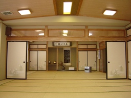 床の間に掛け軸や花瓶が飾られ、右端にストーブが置かれている24畳の日本間講習室を隣の集会室側から撮影した写真