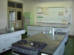 前方に冷蔵庫と掲示物が貼られたホワイトボードがあり、2口ガスコンロが設置されている調理台が2つある調理実習室の写真