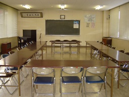 前方にピアノやスタンドマイク、黒板があり、長机とパイプ椅子がコの字型に設置されている農事研修室の写真