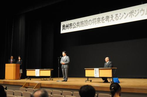 「公共施設の将来を考えるシンポジウム」と書かれた横断幕が会場の舞台上に掲示され、 小沢市長が舞台上のスタンドマイクの前に立ち、挨拶している様子の写真