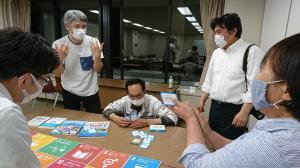 若生 和江代表が1枚のカードを持ち、花澤 淳氏が机にカードを手元に並べ、松本 崇氏が身振り手振りで説明をしている白熱した4名のトークを聞いている男性2名の写真