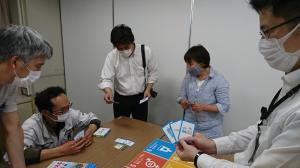 5名の人達が机の周りでカードを手に取り眺めている写真