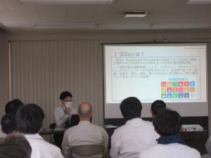 前方のスクリーンに「SDGs」に関する資料が映しだされ、スクリーンの横に座り講演をしている男性、職員の方々が座って話を聞いている様子を後方より写した写真