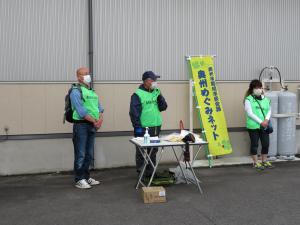 屋外の建物の前に蛍光色の緑色のベストを着た3名の参加者が間を開けて立っている写真
