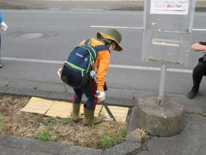 リュックを背負いオレンジ色の洋服を着た参加者が、バス停横の花壇の中のごみを火ばさみで拾っている後ろ姿の写真