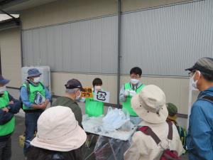 集められたゴミの周りに参加者達が集まり、黄緑色のベストを着た1人の方がプラスチックマークが描かれた紙を持ち、もう一人の黄緑色のベストを着た男性が話をしている様子の写真