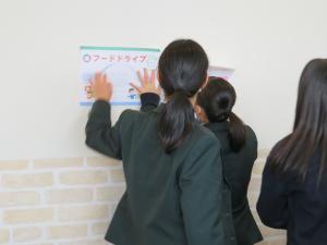 制服姿の女子生徒たちが、白壁に「フードドライブ」と書かれたイラスト付きのポップを貼ろうとしている写真