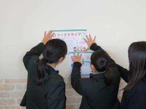 3名の制服姿の女子生徒たちが、白壁に2枚のポップを貼ろうと手で支えている様子の写真