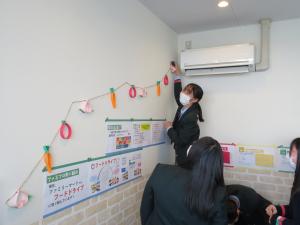 壁に貼られたポップの周りに糸に通した画用紙の飾りを貼り付けている女子生徒の写真