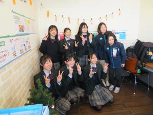 ポップや飾りつけの終わった部屋の一角で、6名の女子生徒と2名の店員が一緒に並び記念撮影をしている写真