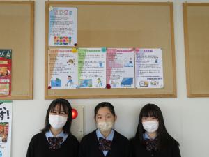 壁のボードに5枚のチラシを並べて展示し、3名の女子生徒と一緒に記念撮影を撮っている写真