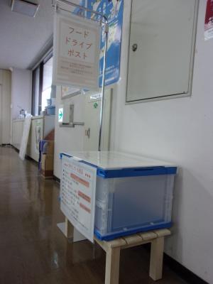 壁際に「フードドライブポスト」を紹介する紙が展示され、木製の台の上に青のクリアボックスが用意されている写真