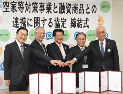 机の上に締結書を広げて並べ、関係者の5名が横に並んで手を重ね合わせて団結を表し、笑顔で写っている協定締結式の写真