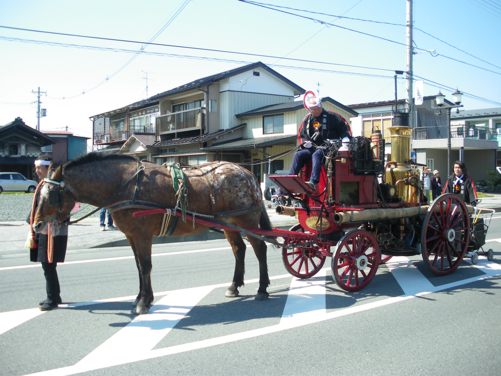 前輪が後輪より小さめに作られた赤色の車体の蒸気ポンプを馬が引いている様子の写真