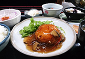キノコのあんかけソースがたっぷりかかった、豆腐ハンバーグと、サラダ、小鉢、漬物、ごはん、汁物と豆乳プリンが黒いお膳に乗った豆太郎セットの写真