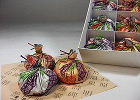 金、銀のベースに紫、緑、オレンジでデザインされた包み紙で、きんちゃく型に包まれた絞り羊羹が、茶色の敷紙の上に3つ並び、その後ろに箱入りの絞り羊羹が置かれている写真