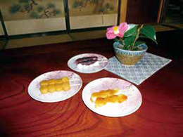 木目調のテーブルに、鉢植えの花と、3種類の団子が2本ずつ盛り付けられた白にピンクの模様が入った丸い皿が3枚並んでいる写真
