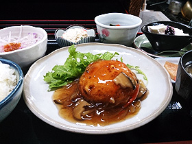 キノコのあんかけソースがたっぷりかかった豆腐ハンバーグと、サラダ、小鉢、漬物、ごはん、汁物と豆乳プリンが黒いお膳に乗った豆太郎セットの写真